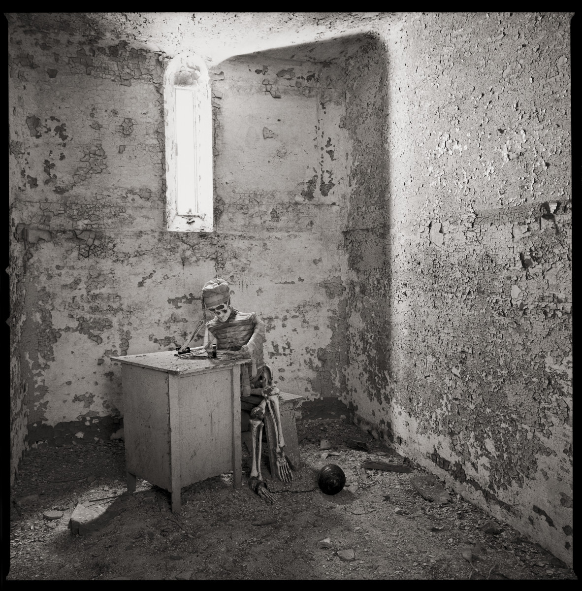 Self-Portrait by Eric T. Kunsman  Image: ID: A black and white image shows room with a window against the back wall and a desk bellow the window.  The paint is peeling and there is dirt and debris onto floor.  There is a skeleton sitting at the desk with prisoner clothing on.  