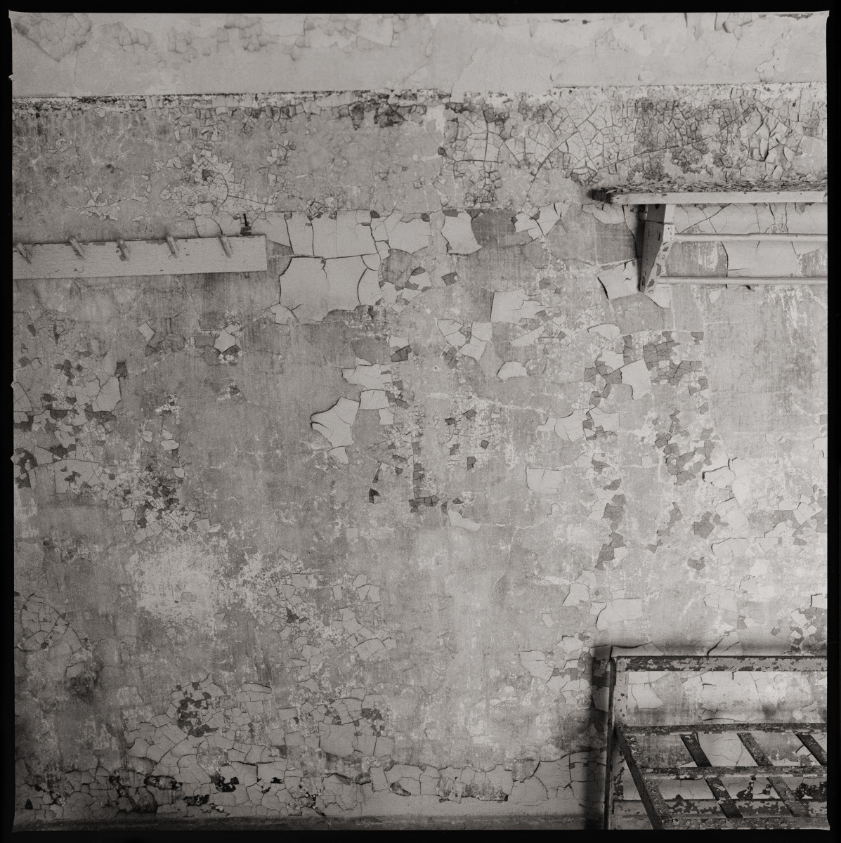 Solitude by Eric T. Kunsman  Image:  ID: A black and white image shows a wall with the paint peeling and cracking cement.  There is a corner of a metal bed frame in the lower right corner.