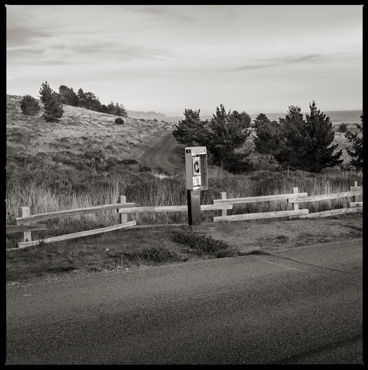 Unknown Number– Point Reyes National Seashore, CA by Eric T. Kunsman  Image: ID: A black and white image shows a payphone in front of a wooden fence.  Behind the fence is a grassy hill with trees.