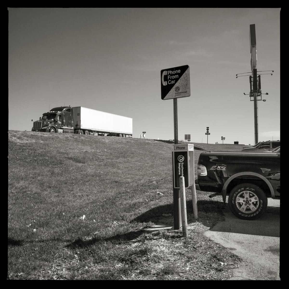 Unknown Number- Outside Springfield, NY by Eric T. Kunsman  Image: ID: A black and white image shows a payphone at the corner of a parking lot with a sign that reads "phone from car".  There is a semi truck in the background on the road.