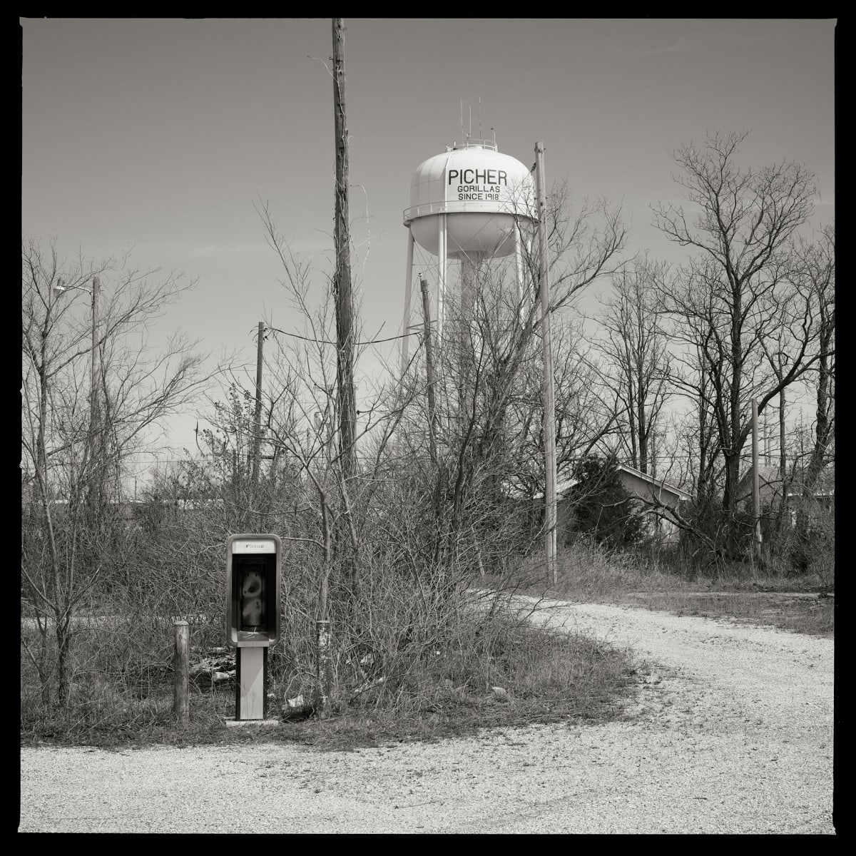 Unknown Number- Superfund Site, Picher, OK by Eric T. Kunsman  Image: ID: A black and white image shows a gravel path that loops around to the left. There is a payphone in just behind the path on the left.  There is a water tower to the right of the path further back.