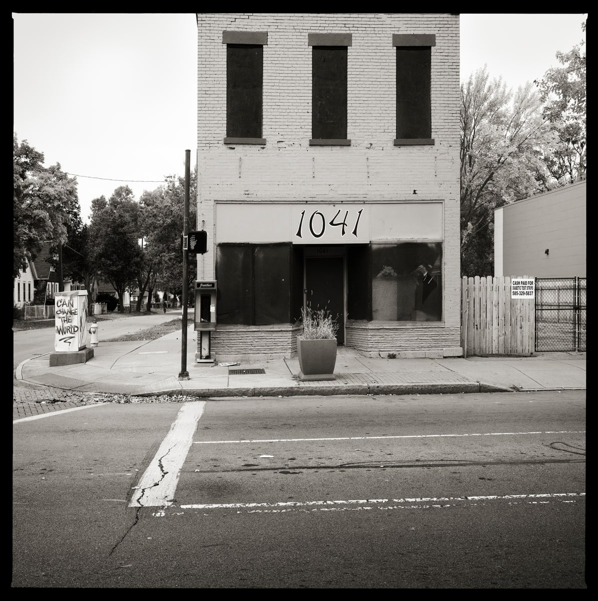 Unknown Number – 1041 North Clinton Avenue, Rochester, NY 14621 by Eric T. Kunsman  Image: ID: A black and white image shows a rectangular building that is closed.  The building is on a corner of a street.  There is a payphone located on the left side of the building's face.  There is also a potted plant in front of the building.