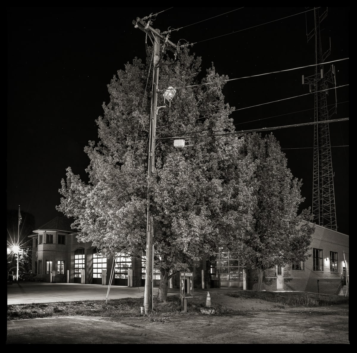 Unknown Number – Brighton Fire Department, 3100 East Avenue, Brighton, NY 14610 by Eric T. Kunsman  Image: ID: A black and white image shows a group of trees at night.  Behind the trees is a building that is lit up.  Under the closest tree is a payphone.  