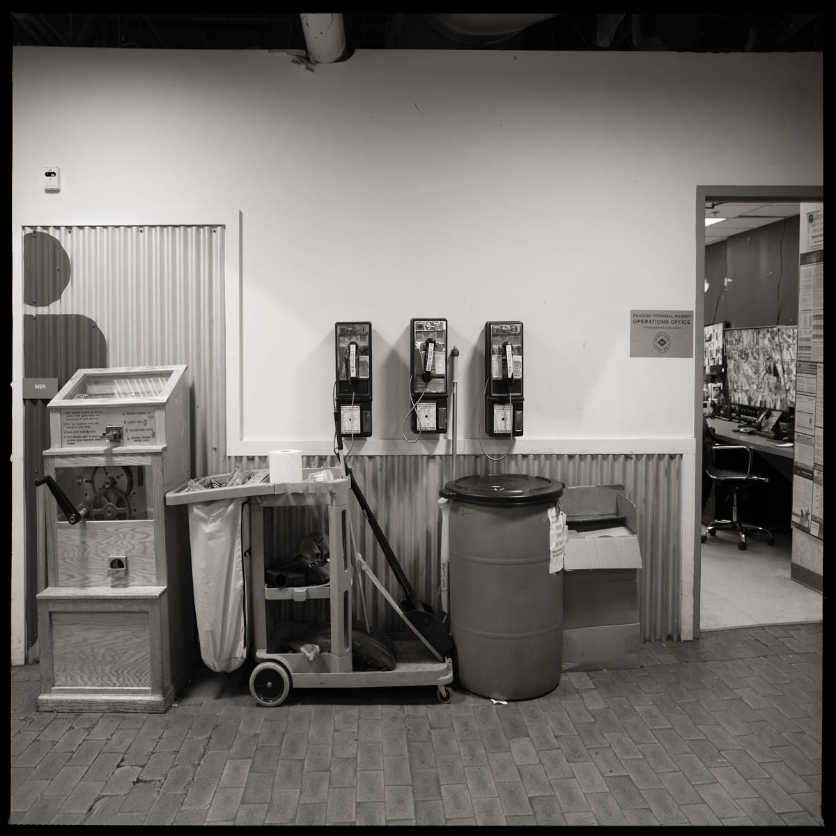 Unknown Numbers- Reading Terminal Market, Philadelphia, PA by Eric T. Kunsman  Image: ID: A black and white image shows three pay phones mounted on a wall.  In front of the payphones are trash cans and a cleaning cart.  