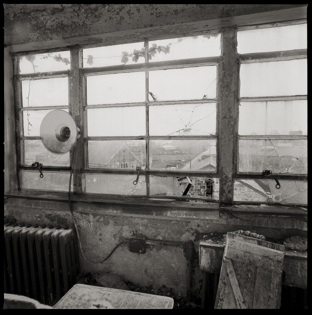 View from the Guard Tower by Eric T. Kunsman  Image: ID: A black and white image shows a view through a cracked window.  In front of the window is an old radiator.  Beyond the window there are buildings visible.