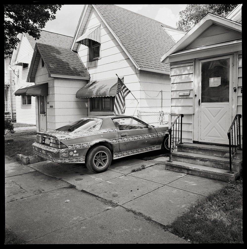 Buffalo, NY by Eric T. Kunsman  Image: ID: A black and white image shows a car with lines of "Sunoco" stickers around the car.  It is parked in a residential area.