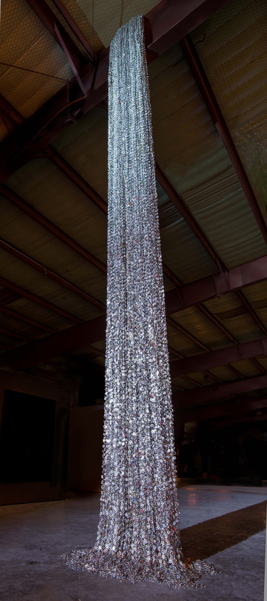 Cascade: collective thirst by Virginia Fleck  Image: Cascade: collective thirst is a 27 ft long 
suspended sculpture made form 85K post-consumer aluminum tabs, safety pins and key 
rings. Aluminum can-tabs chained together with safety pins are layered and massed to 
create a dense sparkling texture. The long chains of can-tabs, reflective, glinting in the 
light, awaken a sense of wonder in those who pass by. When a viewer draws near, 
closer inspection reveals that the waterfall is made from tens of thousands of aluminum 
can-tabs, one of many common disposable items that continuously pass through our 
hands. 