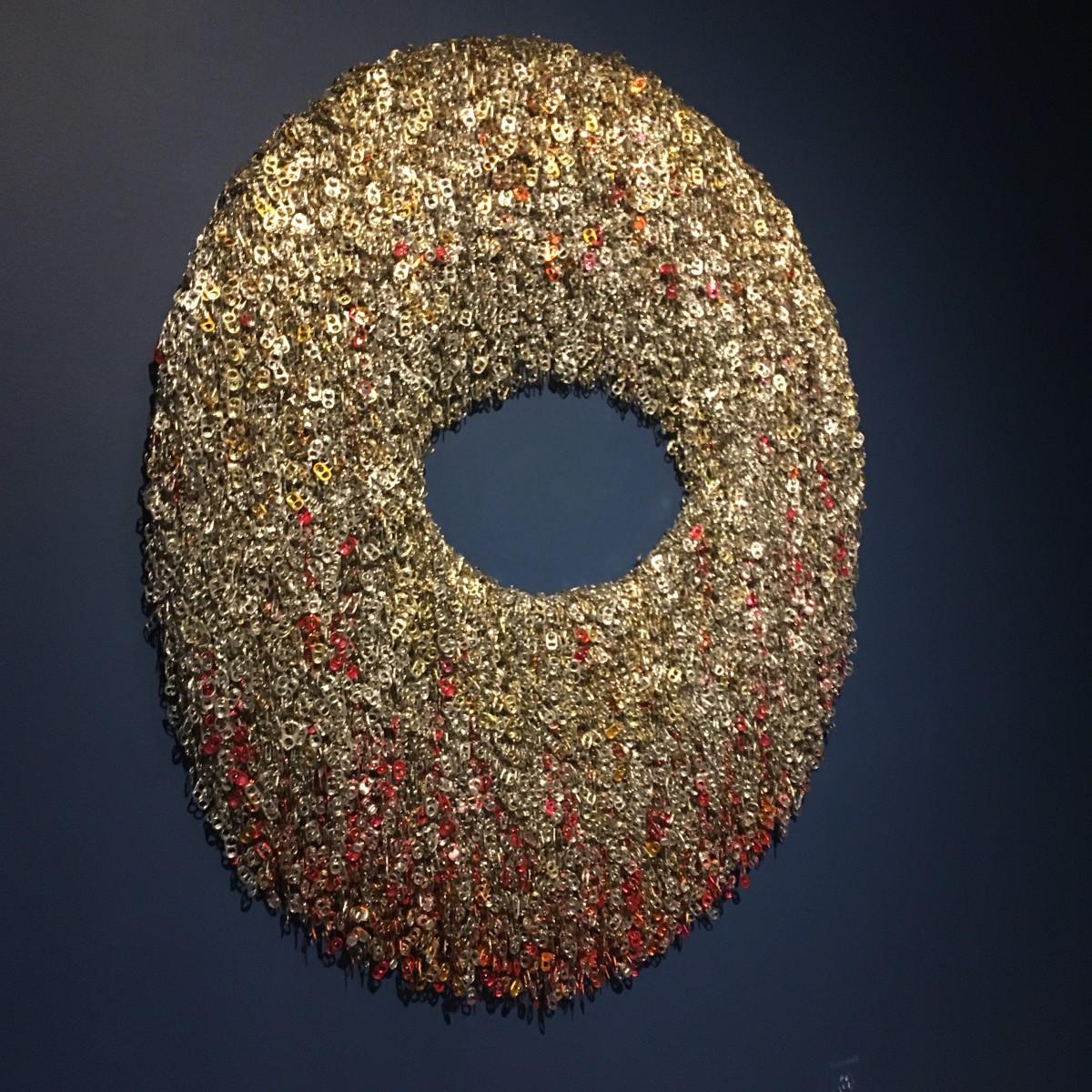 Treasure 2.7 (open center, red striations) by Virginia Fleck  Image: About Treasure Series : Aluminum can-tabs chained together with safety pins are layered, massed and attached to an MDF backing creating a dense sparkling texture. The can-tabs are reflective, glinting in the light, catching the eye of those who pass by them. These shield-like, glittering wall pieces have the presence of valuable treasure, but closer inspection reveals that the treasure is made from can-tabs, one of many common disposable items that continuously pass through our hands. Hangs flush to wall on meatal cleat (included) weight aprox 25 lbs