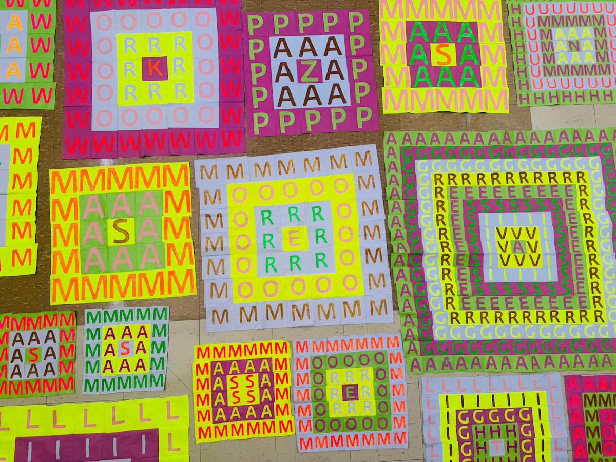 We are looking down at a group of cloth squares lying on the floor. Each square is filled with a grid painted with vibrant contrasting colors. Each square in each grid contains a hand-painted letter. Together they spell out words in English and Spanish that run from the outer edges of the squares to the centers. 