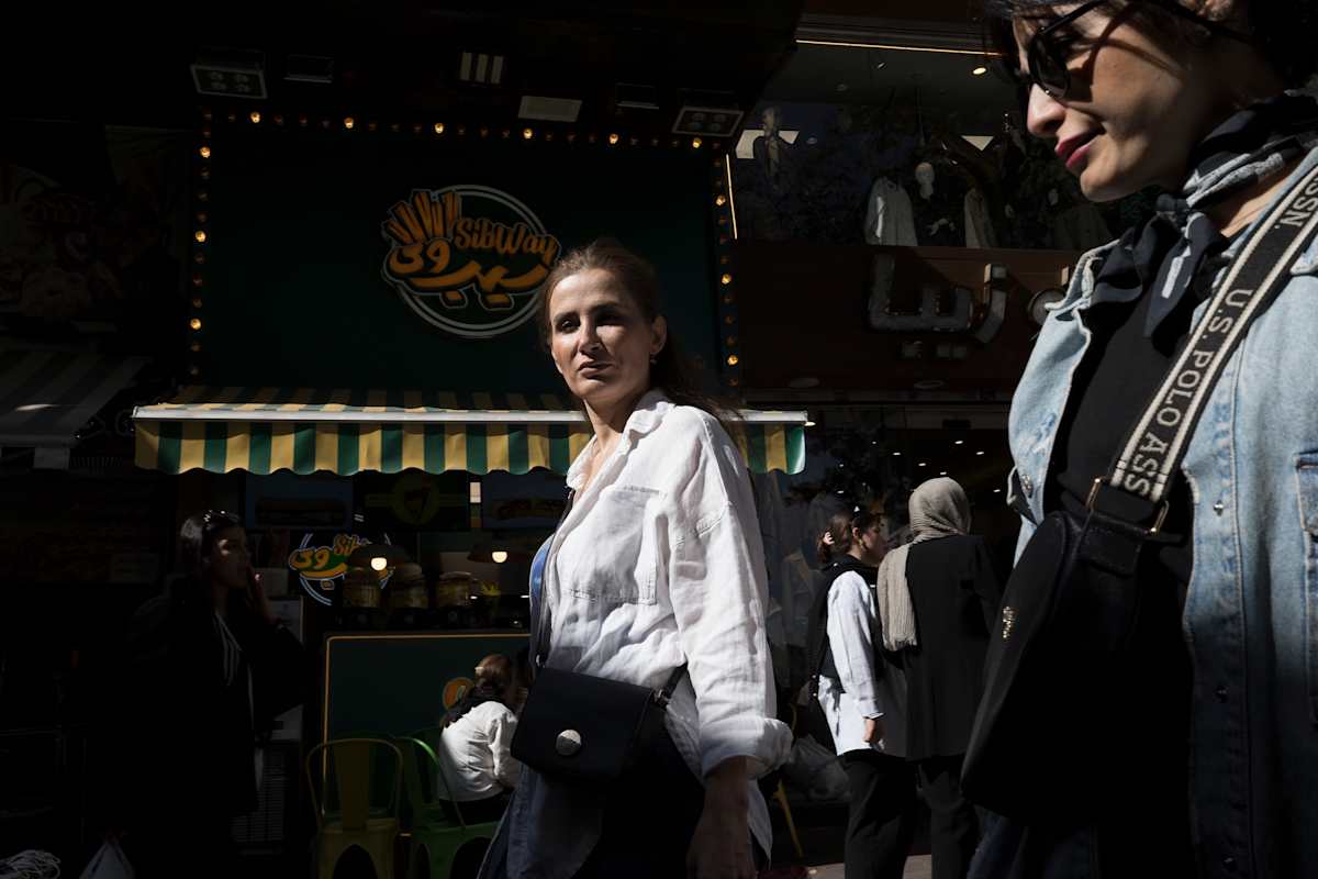 Groups of women are walking past stores on a city street. The name of a business on a building behind them is written in both English and Farsi alphabets. One woman turns in a shaft of light to look at the viewer with an ambiguous expression. She is dressed in smart casual clothes. One woman in the background is wearing a gray hijab, but most of the women in the crowd are bareheaded. 