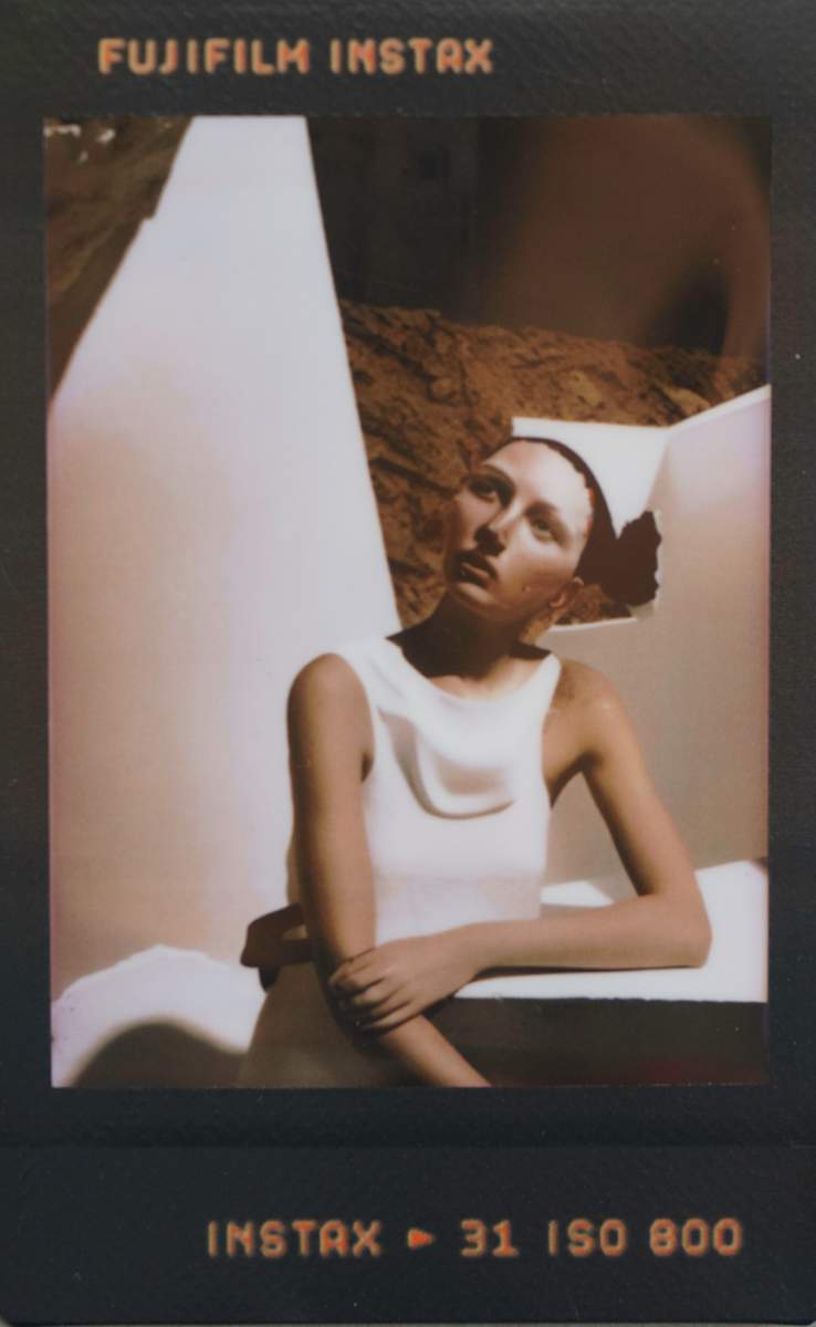 Stand-In Saint / Holding Pattern by Maria Lankina  Image: A standing figure in a pale structured dress holds still against a dark ground, reading like a temporary saint waiting to be swapped out.