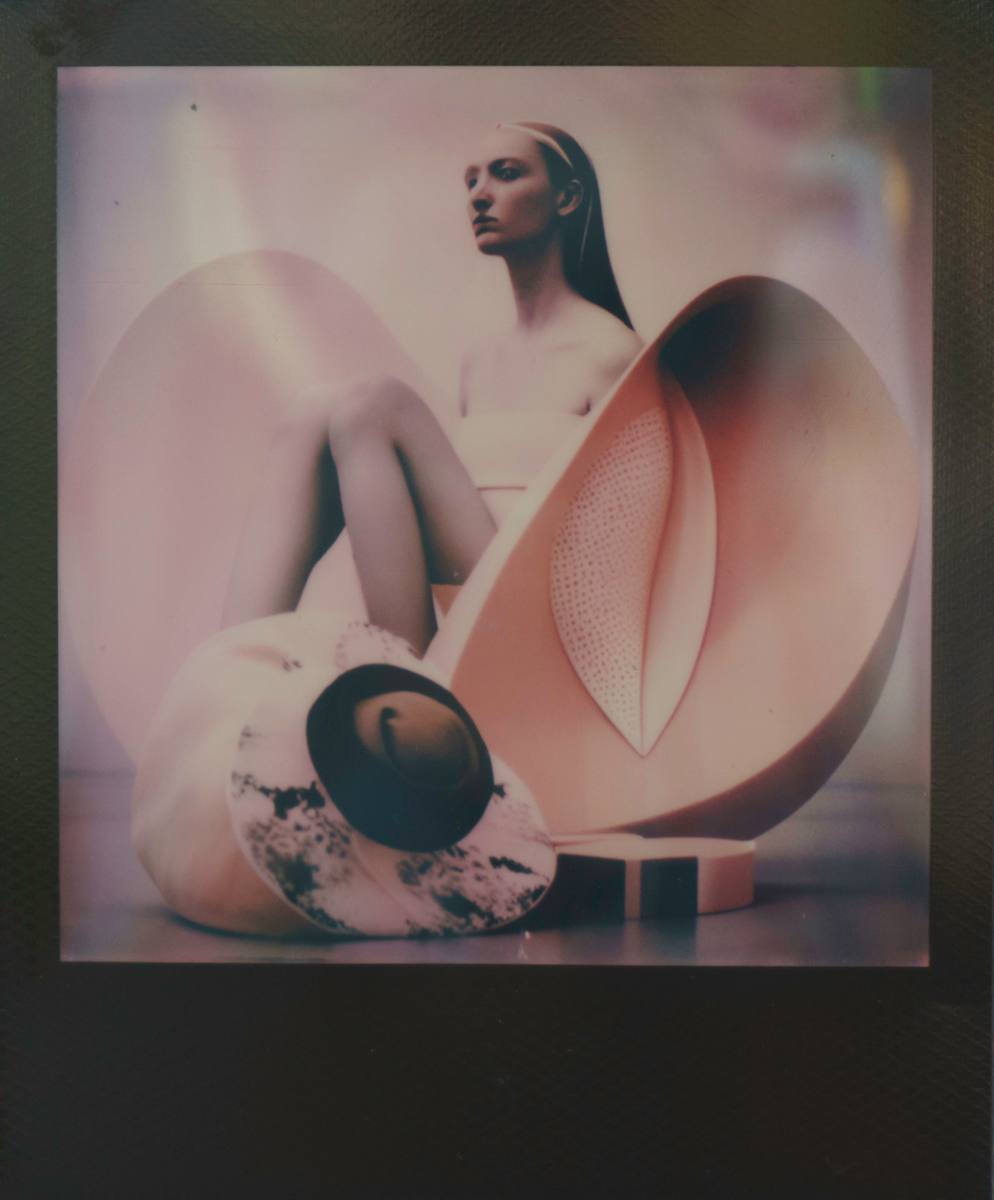 Lily’s Throne / Emergence Chamber by Maria Lankina  Image: A pale figure seated in a peach sculptural shell, part floral throne and part device, held in the soft blur of instant film.