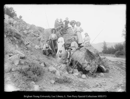 [Chalk Creek Family Fishing] by George Beard  Image: Men, women, and children gathered on large rocks with fishing poles, creek to the right.