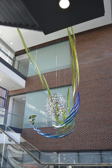 Ondine by Jonathan Clowes  Image: Alt text long description: The image shows a modern interior space with a large hanging sculpture as the focal point. This sculpture is suspended within a multi-story atrium characterized by its brick walls and glass balcony railings. The artwork consists of a spherical core made up of small, reflective pieces that catch the light. Surrounding the core are blue and green curved metallic rods, giving the piece a sense of motion and fluidity. The sculpture hangs from several thin wires attached to the ceiling. A stairway with glass railings is visible in the lower part of the image, ascending to the upper floors. The overall atmosphere is modern and well-lit, with natural light streaming in from unseen sources. Note: the alt text was written with support from the ASU Image Accessibility Creator AI tool. 