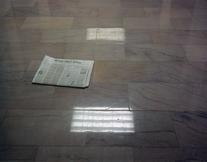 Wall Street Journal, Equitable Building, NYC 2005 by Lisa Kereszi  Image: Alt text long description: The photograph shows a single newspaper lying on a smooth, reflective marble floor. The marble is composed of light gray tiles with subtle, irregular veining in darker gray tones, creating a soft, polished appearance. The newspaper is unfolded, displaying its front page and showing images and columns of text. The tiles are arranged in a staggered pattern, and two rectangular reflections of ceiling lights are visible on the floor, one directly above the newspaper and another near the bottom of the image. Note: the alt text was written with support from the ASU Image Accessibility Creator AI tool. 