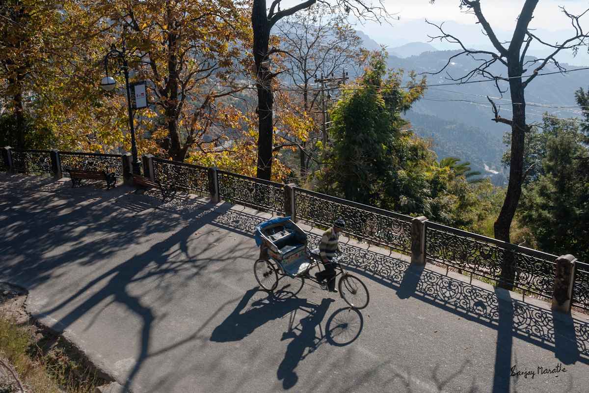 Morning Shadows, Mussoorie (2005) by Sanjay Marathe  Image: I captured this photograph on a winter morning in Mussoorie, Uttarakhand. While exploring the terrain, I found a vantage point overlooking the Mall Road and the hills and waited. Many subjects passed by, but the scene felt incomplete until a rickshaw puller, straining uphill, entered the frame. I clicked the shutter at just the right moment, with no traffic to disturb the composition. The low winter sun cast long, crisp shadows of the ornate wrought iron railing, the tree branches, and the rickshaw, while the soft backlight gave a highlight to the leaves, enhancing the scene’s serene atmosphere.