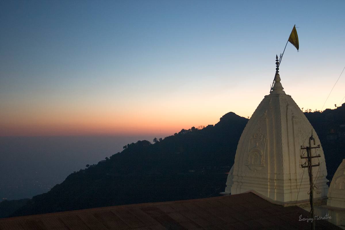 Temple At Dusk, Mussoorie (2005) by Sanjay Marathe  Image: I captured this serene moment just after sunset in Mussoorie, Uttarakhand. The temple stands prominently against the mountain's silhouette, set against a sky that transitions from warm sunset hues to deep blue. A flag flutters gently in the evening breeze, while rolling hills and distant trees add depth to the tranquil backdrop.