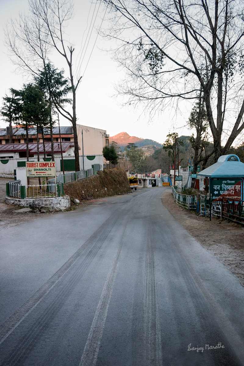 Icy Road, Dhanolti, Uttarakhand (2005) by Sanjay Marathe  Image: A peaceful road winds through Dhanolti, a quaint town in Uttarakhand, India. The biting cold transformed the morning dew on the road into a thin sheet of ice. A few signs point to a tourist complex and local establishments, while trees line the street, framing a distant hill bathed in the warm glow of the morning sun.