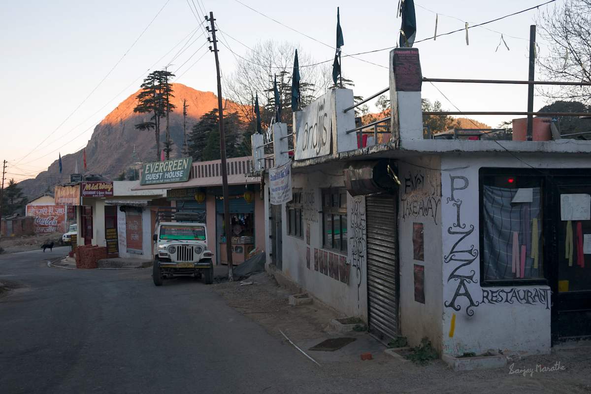 Market, Dhanolti, Uttarakhand (2005) by Sanjay Marathe  Image: An early morning in Dhanolti, Uttarakhand, India, reveals a tranquil street lined with local shops, a guest house, and a pizzeria, with a transport vehicle parked nearby. At the far end, a black dog stands in the middle of the empty road, as if guarding its territory. In the background, a majestic mountain basks in the warm glow of the rising sun, adding to the serene, rural ambiance.
