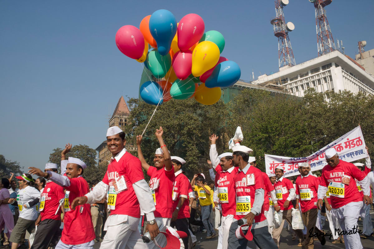 'Mumbai Dabbawalas At The Marathon' (2006) by Sanjay Marathe  Image: Smiling members of the world renowned Mumbai 'Dabbawalas' (tiffin carriers) in bright red shirts and the traditional Maharashtra white head cap participate in a Mumbai marathon.