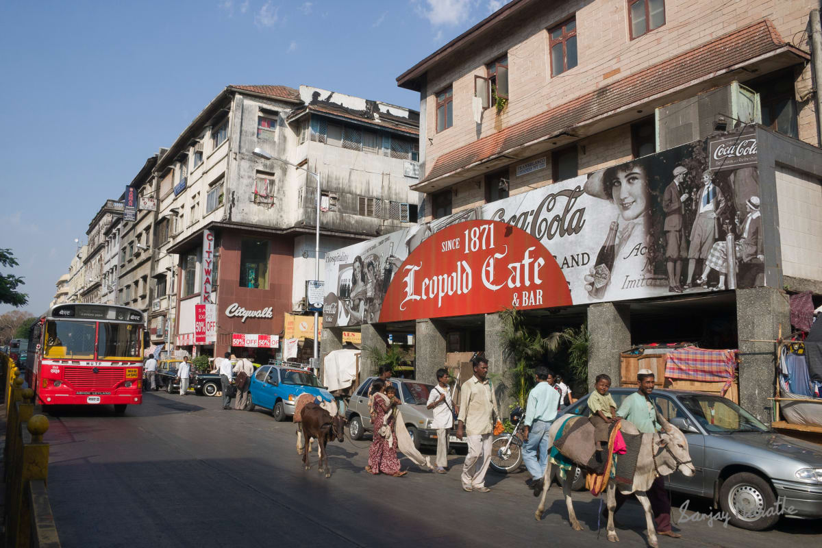 Rural Family Near Leopold Cafe, Mumbai (2006) by Sanjay Marathe  Image: A street performer with  troupe and cows walks past the Leopold Cafe at the Colaba causeway in Mumbai, India.