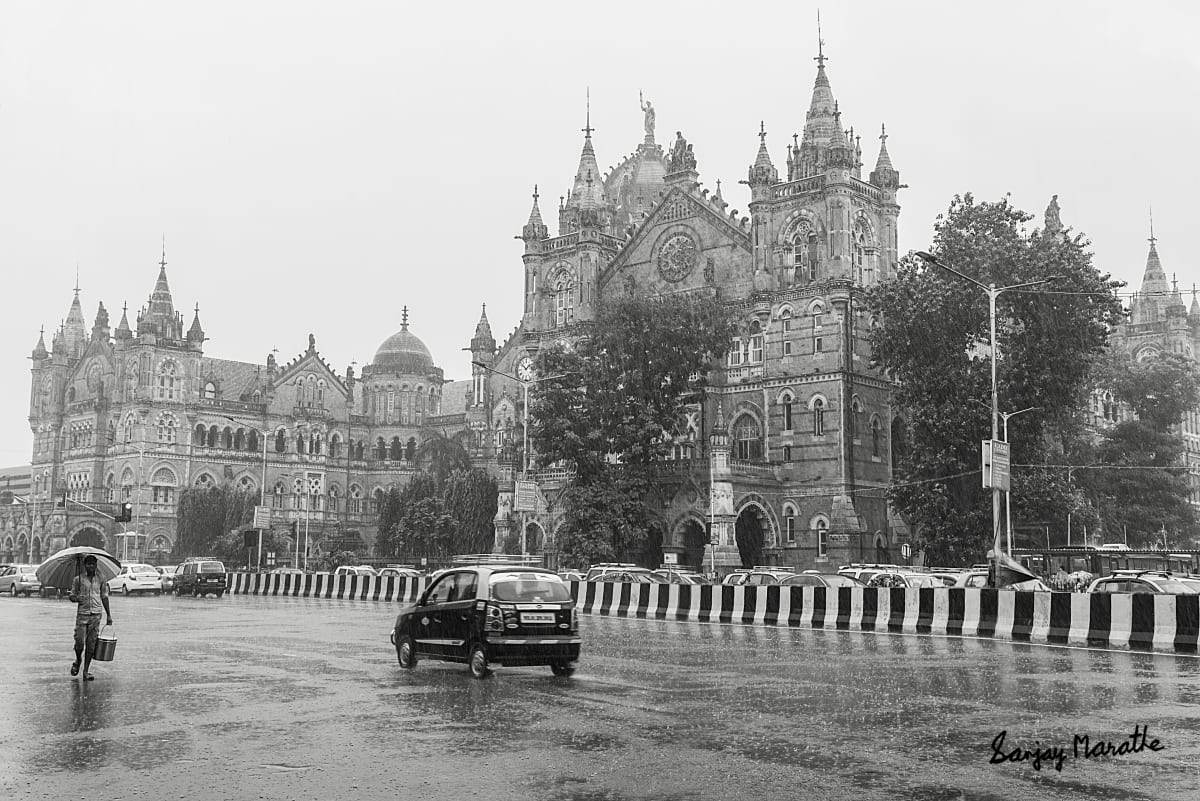 Rain At Victoria Terminus, Mumbai (2019) by Sanjay Marathe  Image: A black and white photograph of Victoria Terminus (now renamed Chattrapati Shivaji Maharaj Terminus) photographed on an unusually rainy day, with sheets of rain pouring down. The minimal traffic resulted in an undisturbed view of the grand building. 

The terminus is a UNESCO world heritage site and a historical landmark, and one of the finest functional railway stations in the world. Built in 1888, it is a magnificent vestige of the British Raj in India. It has an outstanding Victorian Gothic Revival architecture, blended with themes from Indian traditional architecture. It was designed by the British architect F.W. Stevens, who used the stone dome, turrets and pointed arches widely used in traditional Indian palace architecture.