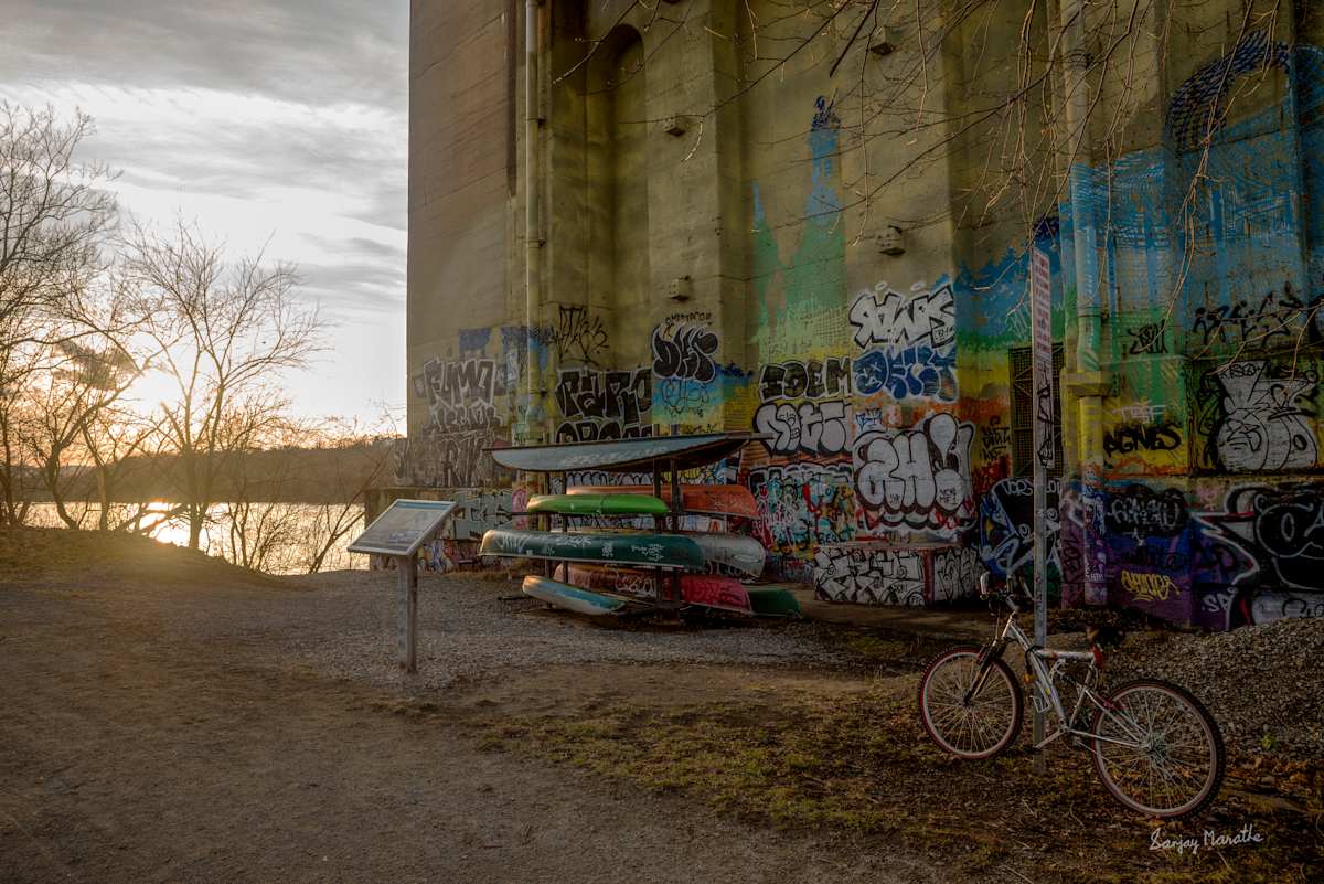 Allegheny River Landscape (2024) by Sanjay Marathe  Image: The setting sun bathed the Allegheny River and the yellow stone bridge column in a warm glow. River rafting boats lay stacked for the night, accompanied by a lone bicycle resting in quiet solitude. I found this serene spot along the Three Rivers Heritage Trail, nestled beneath the Washington Crossing Bridge in Pittsburgh, PA, USA.