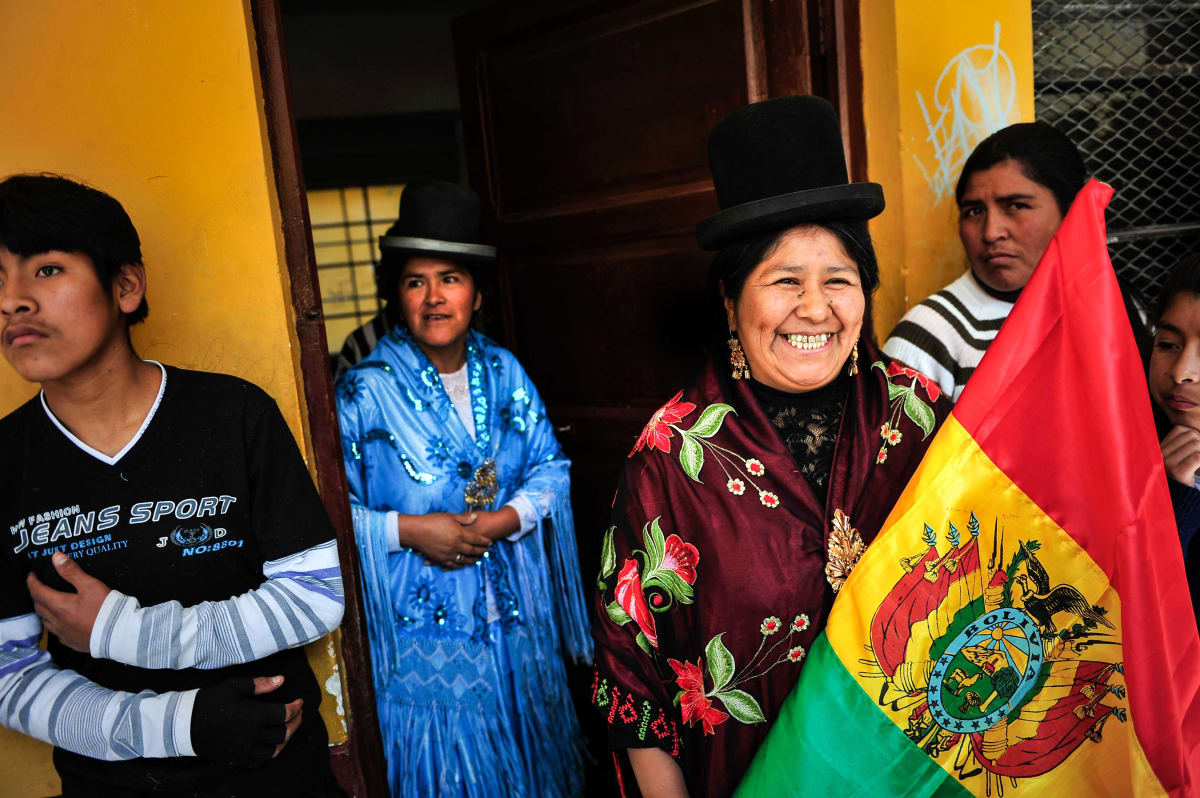 Untitled  Image: Julia and Carmen Rosa wearing their embroidered mantles and cholita hats outside a house, holding the Bolivian flag.