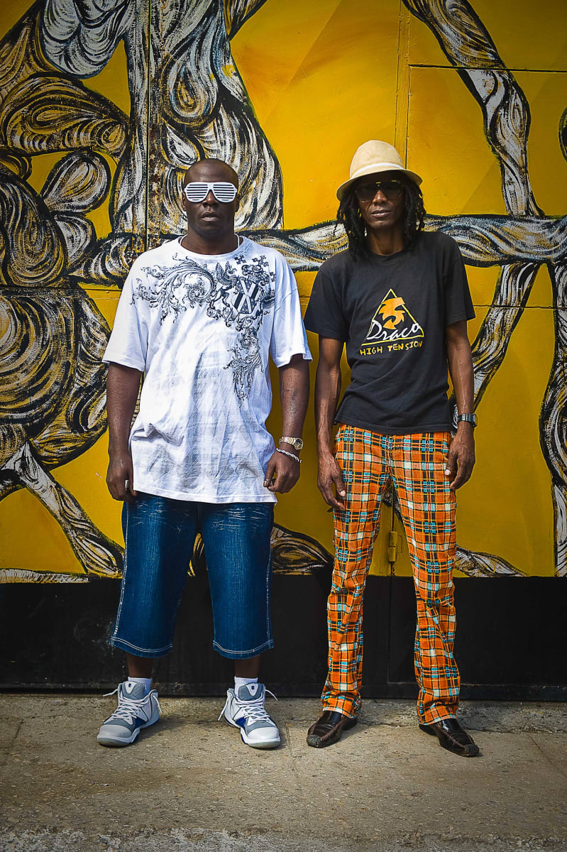Untitled (Two Men)  Image: Two men pose in front of a graffitied yellow wall.