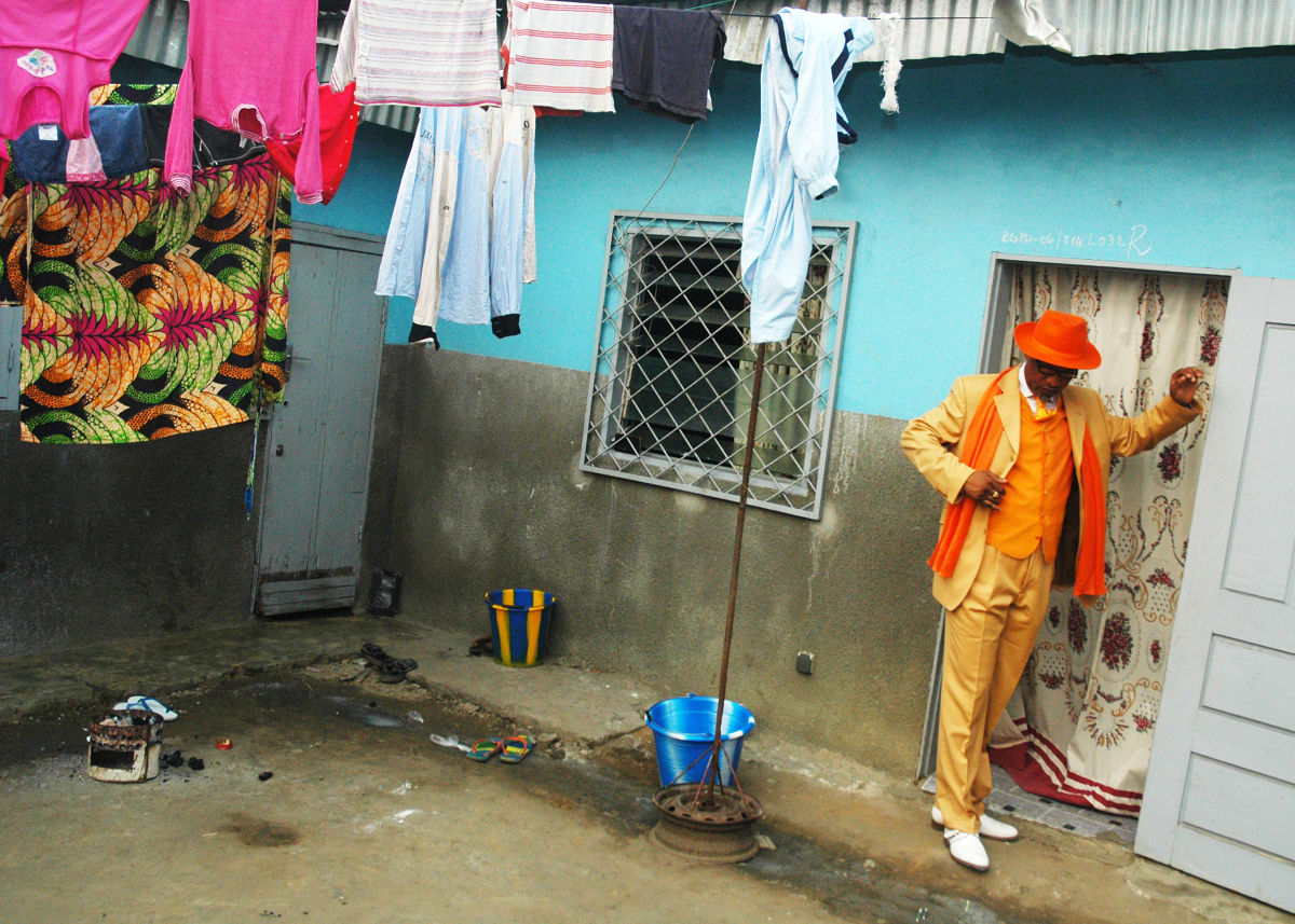 Untitled (Kvv Mouzieto the Sapeur Matsouaniste) by Daniele Tamagni Foundation  Image: Kvv Mouzieto in a bright orange suit topped with matching hat, standing in front of his house. The walls are paited light blue. Over his head, colorful clothes are hanging out to dry. Brazzaville, Congo (2007).