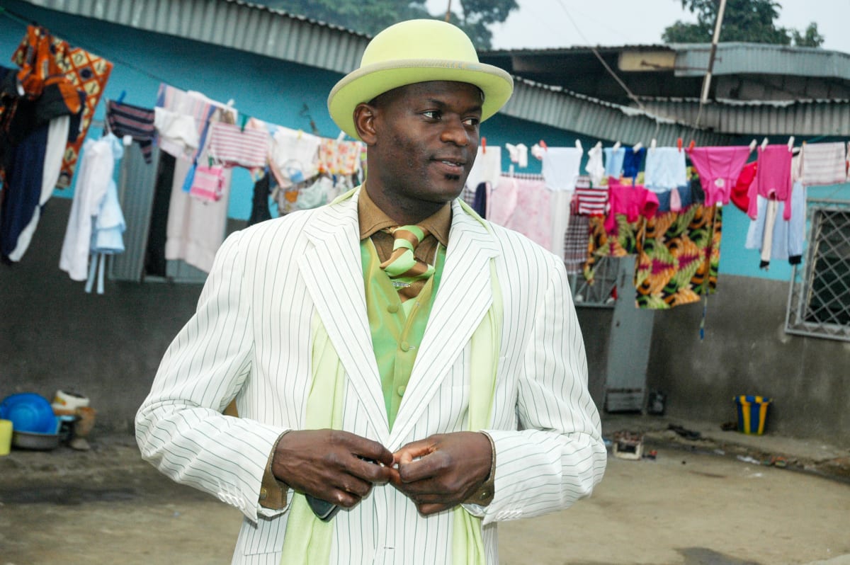 Untitled by Daniele Tamagni Foundation  Image: Kvv Mouzieto's brother, Ange Bienvenu Emmanuel Mouzieto, fastening his suit's buttons outside their family home in Brazzaville. He is wearing a pistachio toned shirt with matching bowler hat, and green and white pinstriped suit. Brazzaville, Congo (2007)