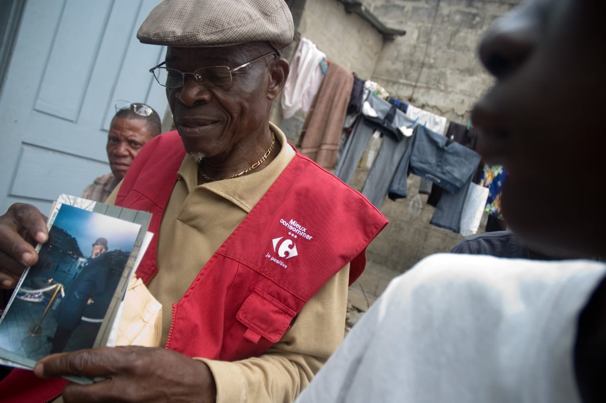 Untitled  Image: Lamame dressed in his daily clothes (beije shirt and red Carrefour gilet), proudly showing some photos of himself in Paris. Lalame, known as 'the old' or 'the Parisian' has a particular passion for Paris, as many Sapeurs do, and has visited the city many times as part of his family is settled there. Very close friend of Kvv, he is particularly well known and respected in Bacongo, where he is sometimes paid to attend weddings or funerals. Brazzaville, Congo (2007)