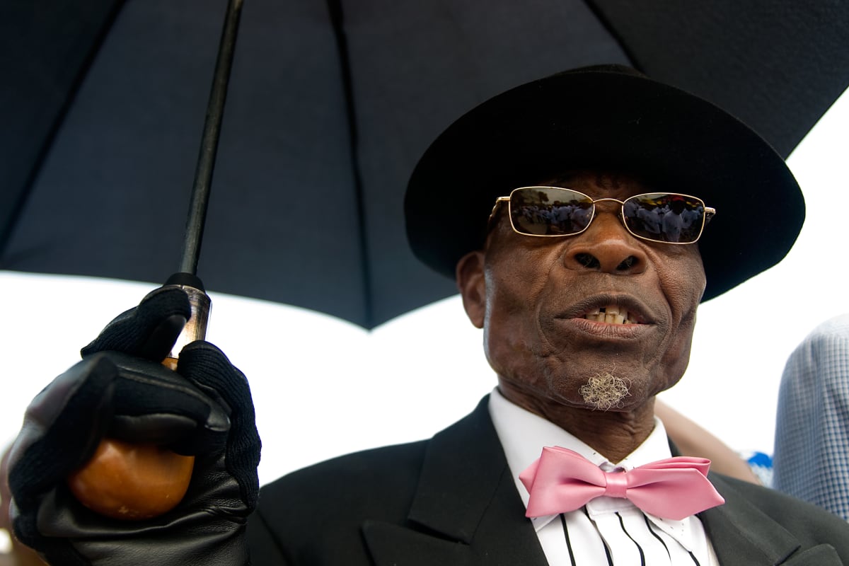 Untitled (Lamame 'the Parisian') by Daniele Tamagni Foundation  Image: Portrait of Lame 'the Parisian' wearing a black suit with matching leather gloves, bowler hat, umbrella and sunglasses, and a white shirt with pink bowtie. Brazzaville, Congo (2007).