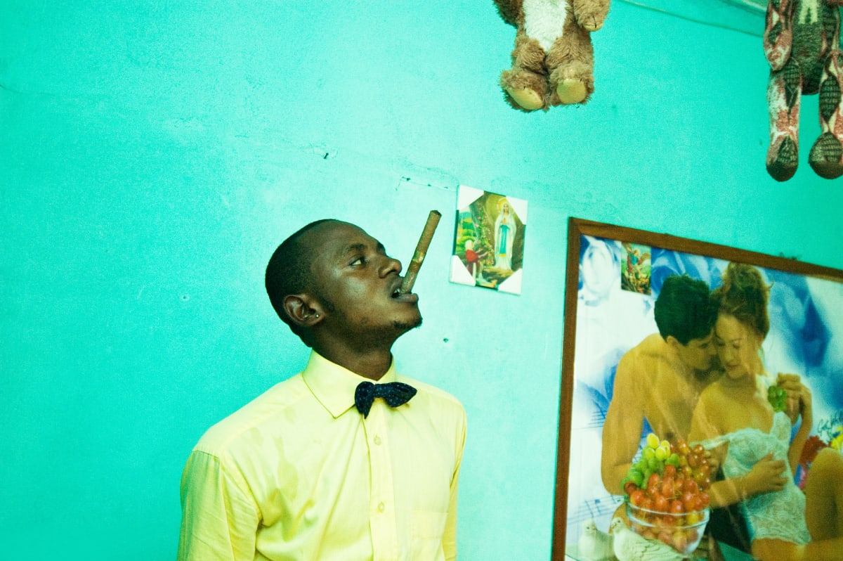 Untitled (Lalhande in his room) by Daniele Tamagni Foundation  Image: Lalhande in his room wearing a yellow shirt and a blue bowtie, biting a cigar. The background wall is light blue, hanging on it an icon of the Virgin Mary and an erotic poster with a caucasian couple. Brazzaville, Congo (2007)