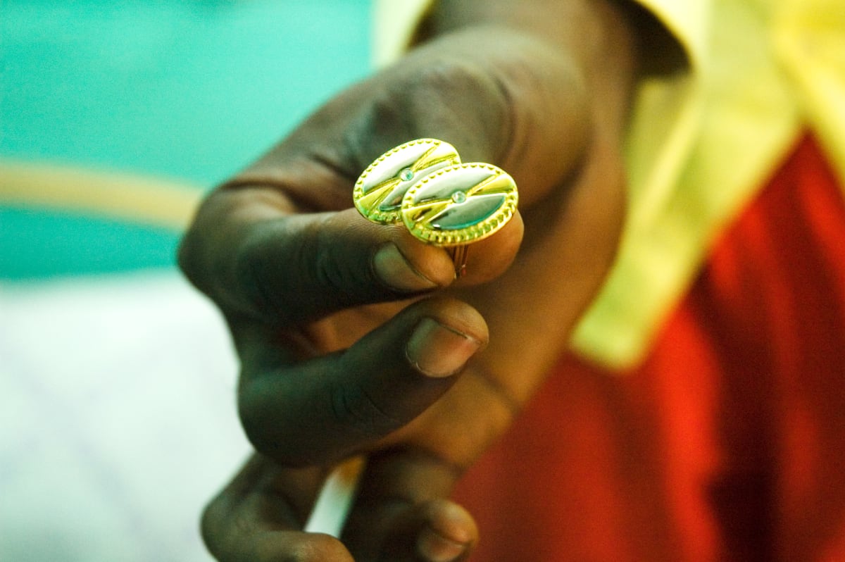 Untitled by Daniele Tamagni Foundation  Image: Close up photograph of Lalhande holding two golden cufflinks. The background, out of focus, shows the tints of the light blue wall and of the yellow and red suit worn by Lalhande. Brazzaville, Congo (2007).
