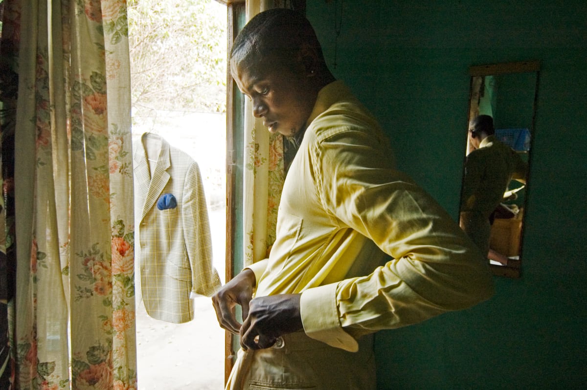 Untitled  Image: Portrait of Lalhande getting dressed inside his house. On his left, the tweed jacket of his suit with a blue pocket handkerchief hangs outside the doorframe. On his right, his reflection in a mirror. Lalhande lives with his mother, loves Italy and is a fan of AC Milan. His grandfather was a Sapeur and a musician of Charanga. Brazzaville, Congo (2007)