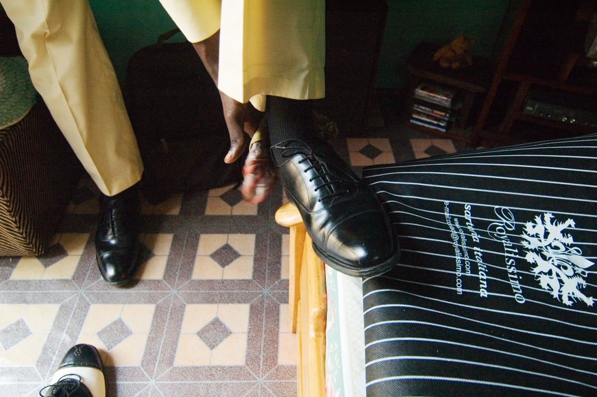 Untitled by Daniele Tamagni Foundation  Image: Lalhande fitting his black leather shoes on the frame of his bed. Besides him, a garment holder with written 'Royalissimo, sartoria italiana' (Royalissimo, italian taylors). Brazzaville, Congo (2007)