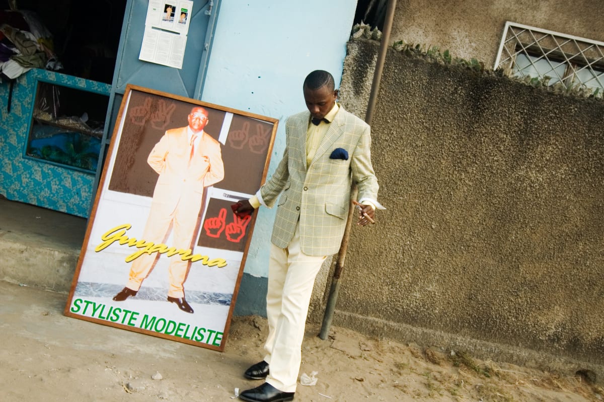 Untitled by Daniele Tamagni Foundation  Image: Lalhande posing for the camera in the streets of Brazzaville, standing next to a framed poster picturing an elegant man and reading 'Styliste Modeliste'. Brazzaville, Congo (2007).