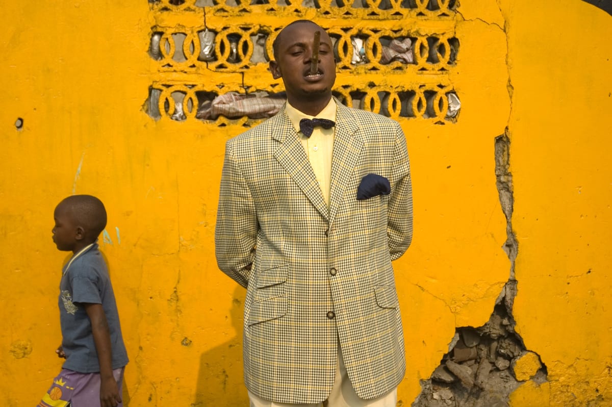 Untitled (Lalhande 'the young') by Daniele Tamagni  Image: Lalhande holding an unlit cigar between his teeth in front of a bright yellow wall with a kid passing by. Brazzaville, Congo (2007)
