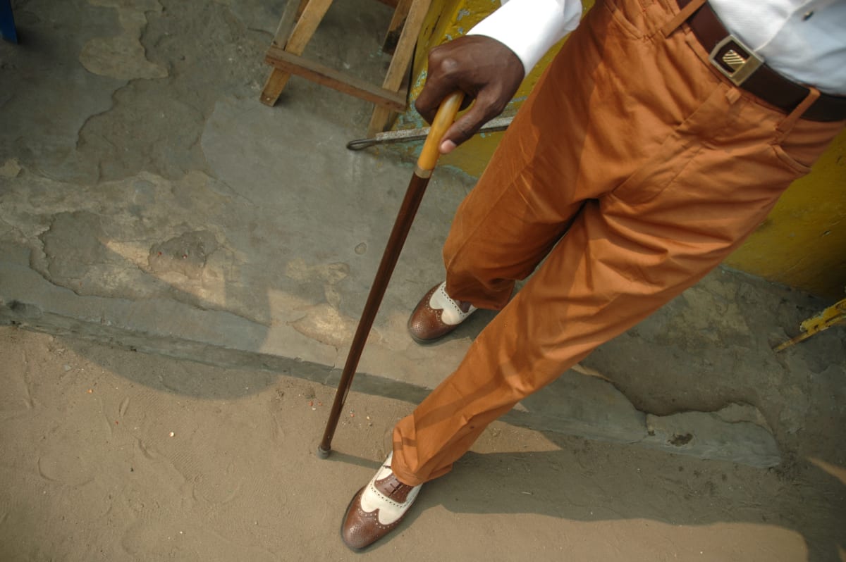 Untitled by Daniele Tamagni Foundation  Image: Bird-eye view of Michel walking down the street with a wooden cane, orange trousers and brown and white leather shoes. Brazzaville, Congo (2007)