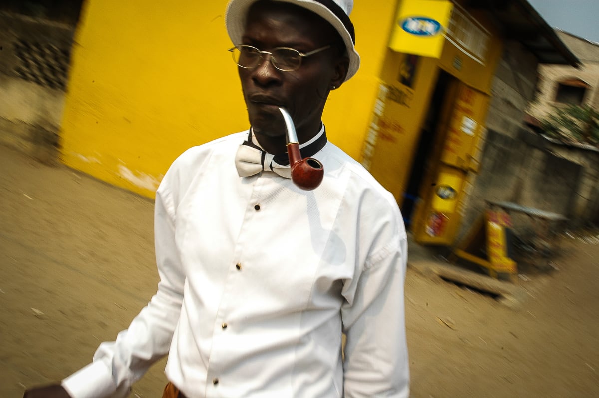 Untitled by Daniele Tamagni Foundation  Image: Michel walking down the street holding his pipe between his lips, with a bright yellow wall as background. Brazzaville, Congo (2007)