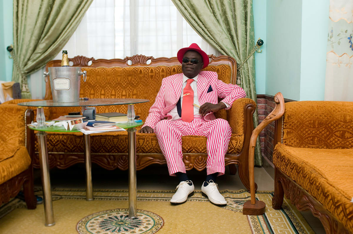 Untitled (Chalereux Abbot) by Daniele Tamagni Foundation  Image: Anselme Badiamo, known by all as the 'abbé Chalereux', wearing a red and pink striped suit and a red hat, sitting on his couch. Brazzaville, Congo (2008)