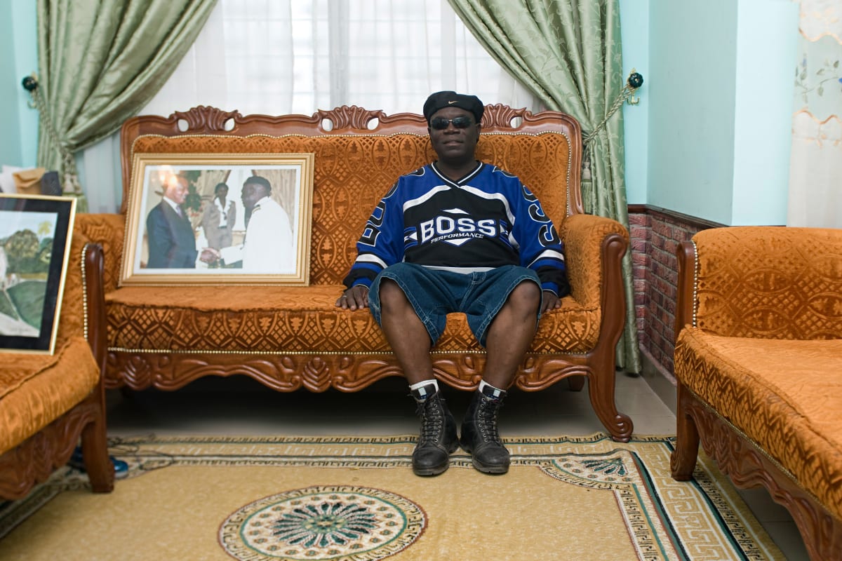 Untitled  Image: Anselme Badiamo posing with a casual 'American' look, sitting next to a photo of the Congolese president. Brazzaville, Congo (2008)