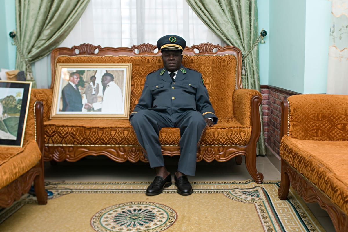 Untitled  Image: Anselme Badiamo, the 'abbé Chalereux', dressed as military chaplain. Anselme doesn't consider himself a Sapeur, but a simple religious man who loves the Sapeur culture because of the values it expresses. As a military chaplain, he has many friends in institutional positions. Here, he sits besides a photograph of the Congolese president. Brazzaville, Congo (2008)