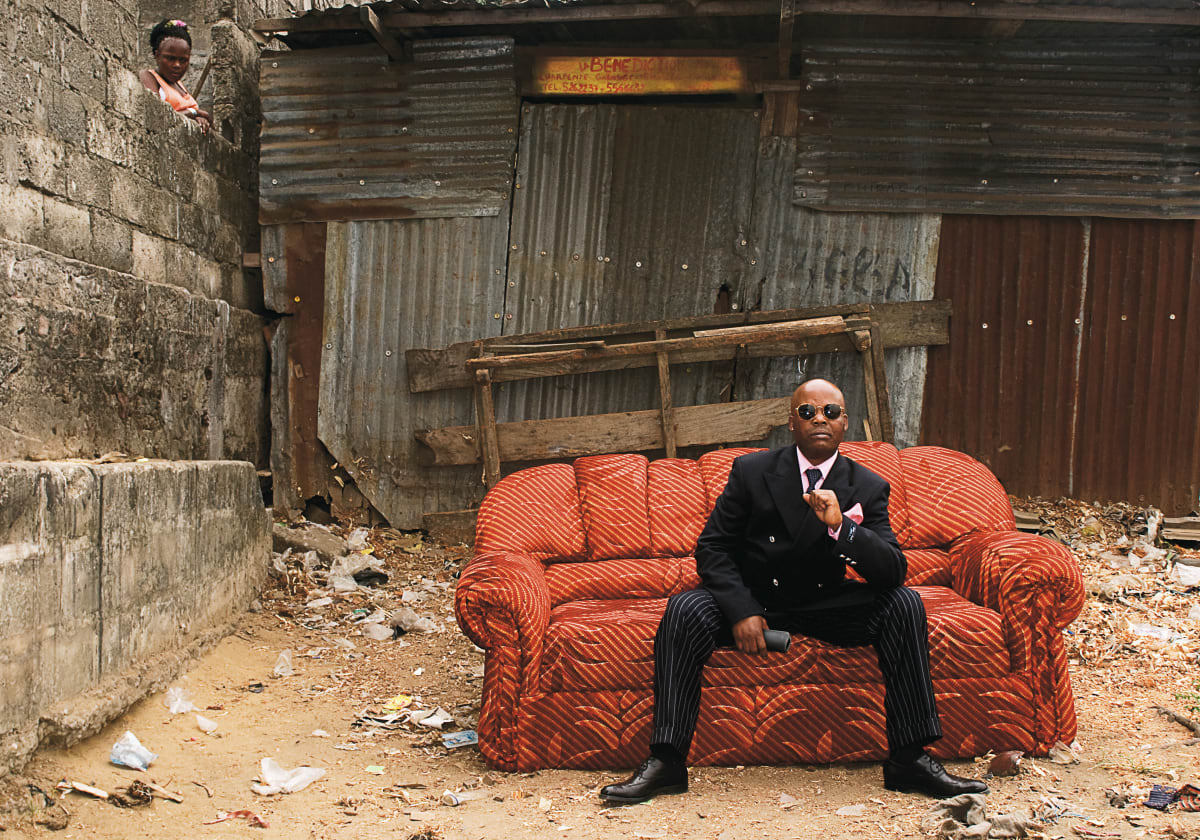 Untitled (Mayembo) by Daniele Tamagni Foundation  Image: Mayembo in a black suit with pink shirt and poket handherchief, sitting on a couch in a street of Brazzaville, Congo (2008)