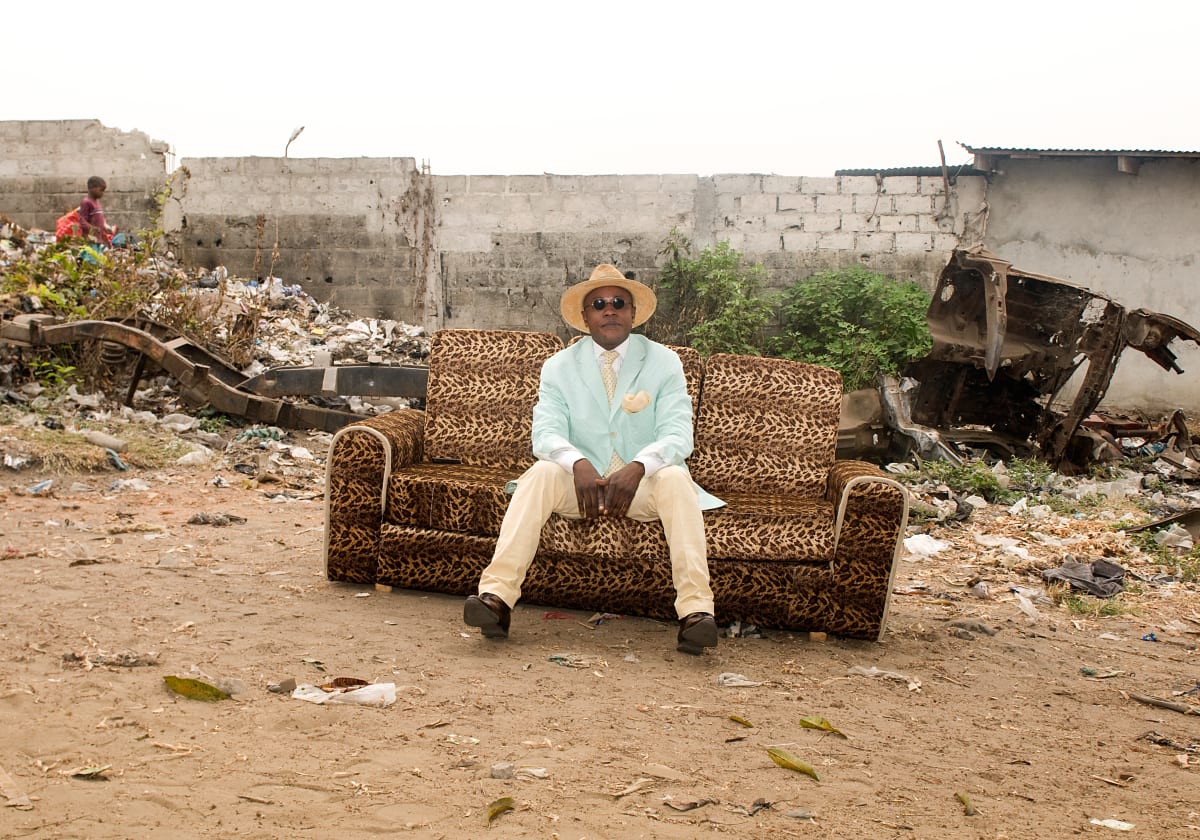 Untitled (Dany)  Image: Dany wearing a light blue jacket paired with creme trousers, tie and handkerchief, sitting on a leopard print couch in an outdoor space. "Sape is the art of creating beautiful harmonies from a combination of colours and styles to bring out the elegance of the dress and the poise of the Sapeur." (Dany) Brazzaville, Congo (2008)