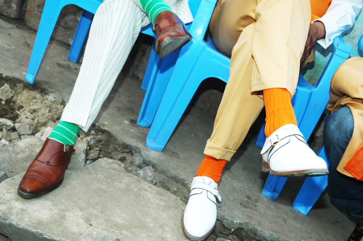 Untitled (Sapeurs Shoes) by Daniele Tamagni Foundation  Image: Photograph of Kvv Mouzieto's and Bienvenu Mouzieto's shoes: brown leather paired with green striped socks on the left and white leather paired with orange socks on the right. The men are sitting on blue plastic chairs. Brazzaville, Congo (2007)