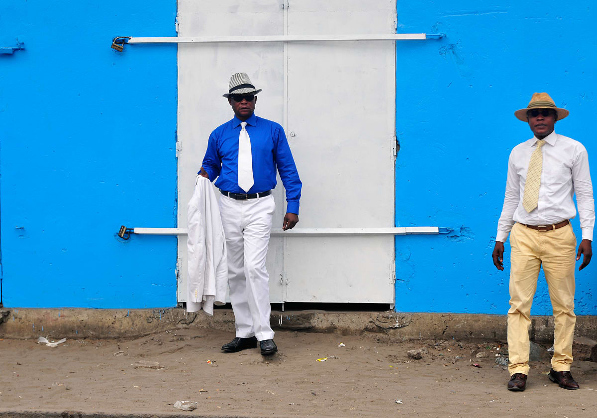 Untitled (The Sapeurs of Moukondo) by Daniele Tamagni Foundation  Image: Dany (right) posing with a friend in front of a blue wall in the district of Moukondo. Brazzaville, Congo (2008)