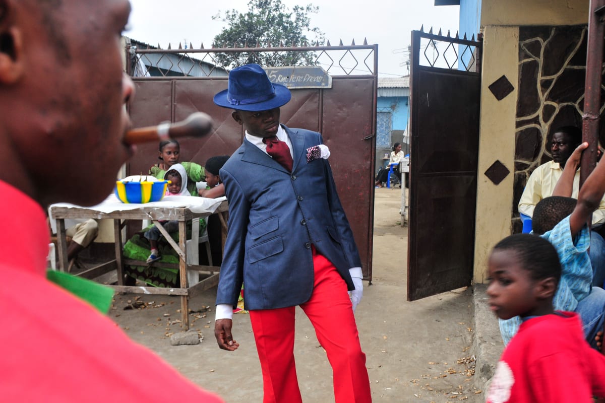 Untitled (Vive la Sape #1) by Daniele Tamagni Foundation  Image: A young Sapeur posing for the camera, wearing red trousers and tie, blue jacket and matching hat. Brazzaville, Congo (2008)