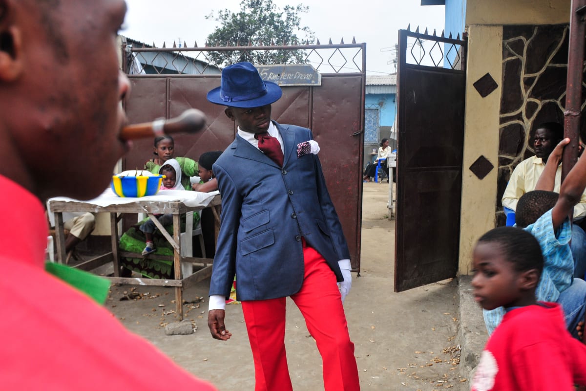 Untitled (Vive la Sape #1)  Image: A young Sapeur posing for the camera, wearing red trousers and tie, blue jacket and matching hat. Brazzaville, Congo (2008)